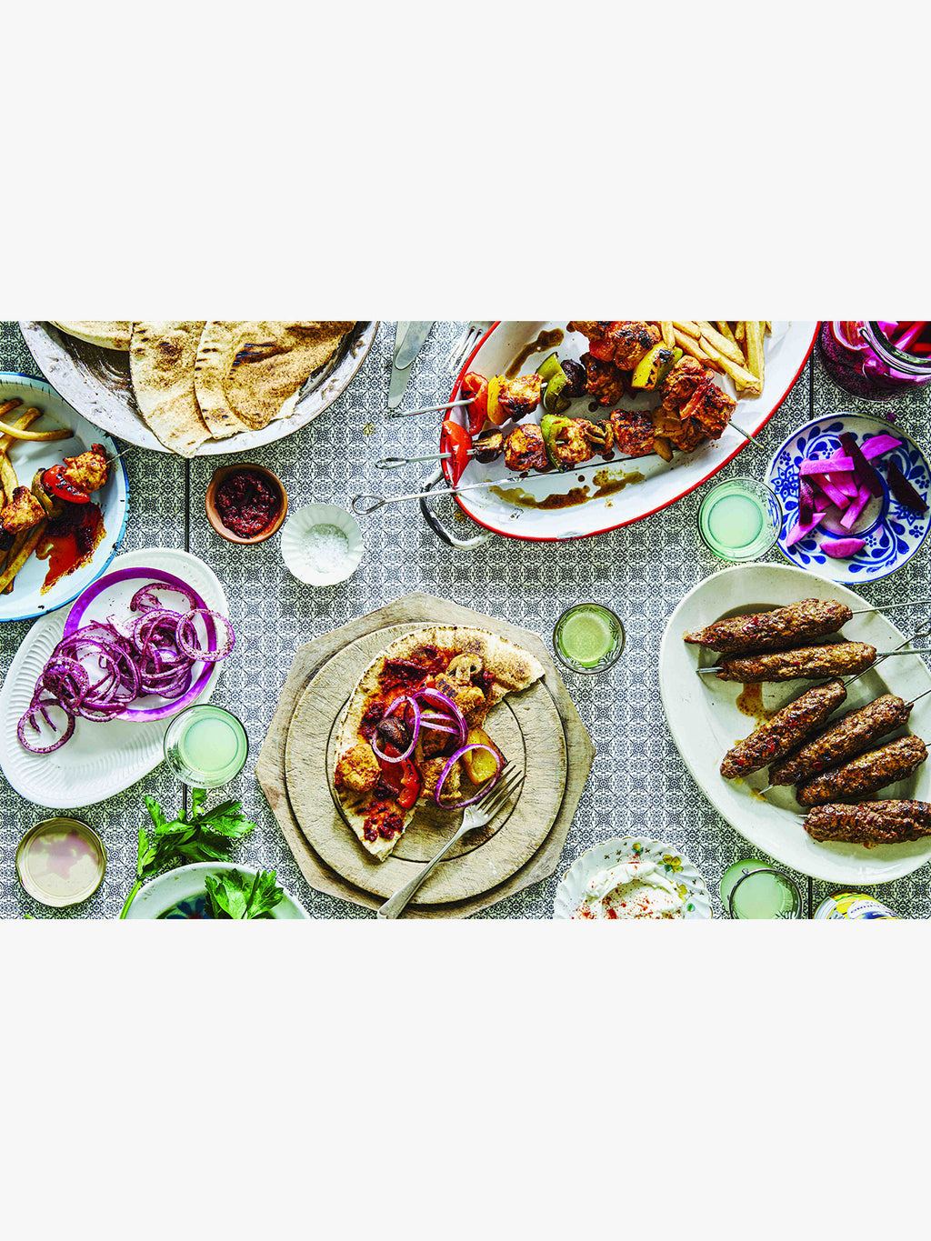 Overhead view of a styled food spread on a patterned tablecloth, featuring plates of grilled meat skewers, flatbread, sliced onions, pickles, and small bowls of condiments.