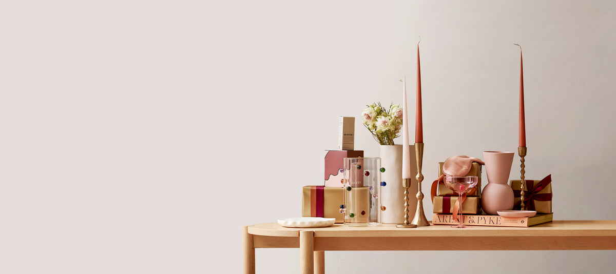A styled tabletop arrangement in soft neutral and pastel tones, featuring wrapped gift boxes, a small vase with dried flowers, tall taper candles, a pink vase, and stacked books on a light wood table against a plain off-white wall.