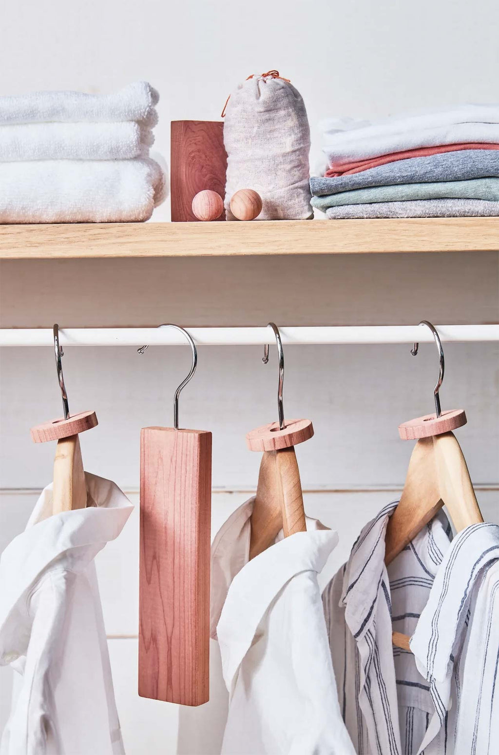 Wooden hangers with round pinkish wooden tops and a rectangular wooden closet organizer block hanging on a white rod in a closet. Folded towels and folded garments are stacked on a wooden shelf above.