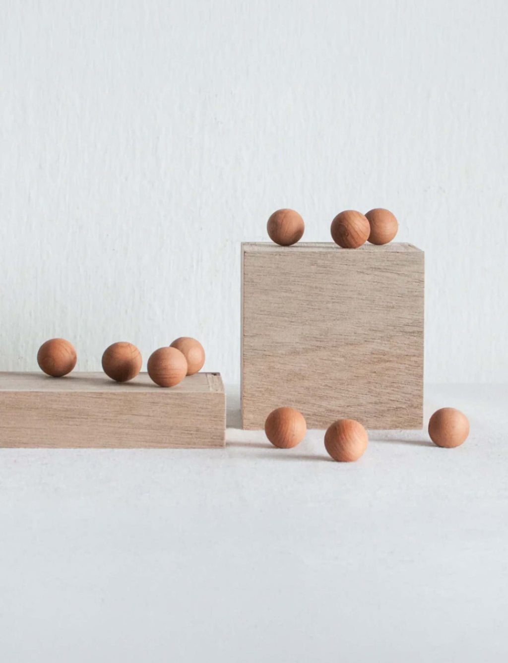 Natural wood blocks of different heights arranged on a light gray surface against a white wall, with small round wooden cedar balls placed on and around them.